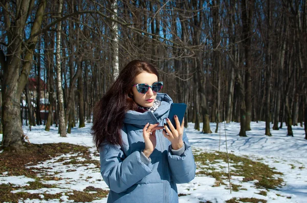 Приваблива жінка в сонцезахисних окулярах і синьому зимовому пальто, використовуючи смартфон, проводячи час на відкритому повітрі в парку в ранній весняний час — стокове фото
