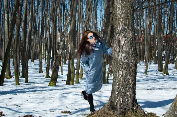 Attraktiv kvinna i solglasögon och blå vinterkappa som står bakom kala träd medan spendera tid utomhus i parken under tidigt våren — Stockfoto