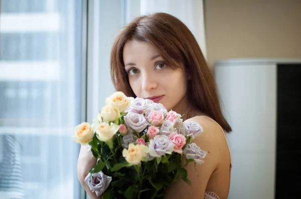 Schönes junges Mädchen, das am Fenster mit einem Blumenstrauß steht und in die Kamera schaut, drinnen Frauenporträt. Warten auf ein Frühjahrskonzept — Stockfoto