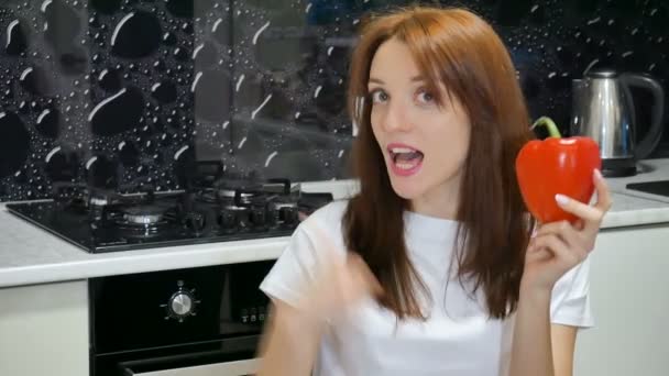 Funny excited young woman with big red bell pepper showing thumb sitting at the kitchen at her home — Stock Video