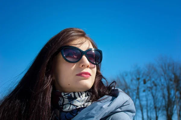 Mulher bonita em óculos de sol na moda e roupas de inverno posando no céu brilhante e árvores nuas . — Fotografia de Stock