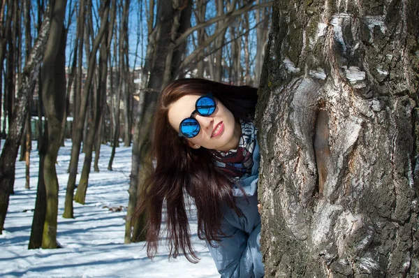 Närbild utomhus porträtt av fantastisk kvinna i solglasögon och blå vinterkappa som står bakom kala träd i parken under tidigt våren — Stockfoto