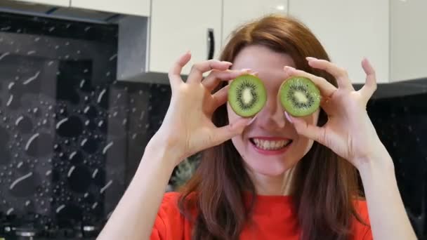 Menina jovem louco mostrando kiwi sobre seus olhos enquanto prepearing comida na cozinha. Conceito de emoções humanas positivas . — Vídeo de Stock