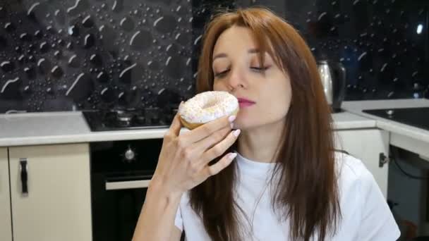 Inomhus porträtt av en nöjd ganska vuxen flicka äter donut sitter vid köket hemma. Välsmakande matkoncept. — Stockvideo