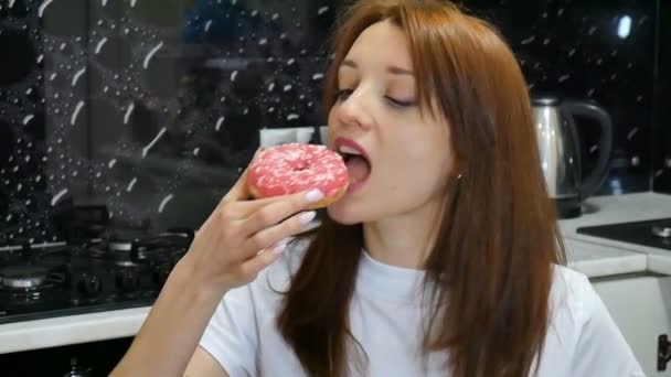 Menina com fome de cabelos longos comendo donut vermelho na cozinha em casa. Conceito de comida saborosa — Vídeo de Stock