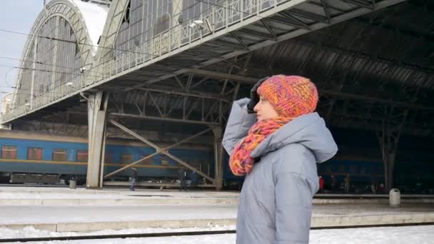 Ritratto di una giovane donna in cappotto blu e cappello colorato alla stazione ferroviaria durante la soleggiata giornata invernale. Concetto di viaggio. Stile di vita, viaggiare, vacanza — Video Stock