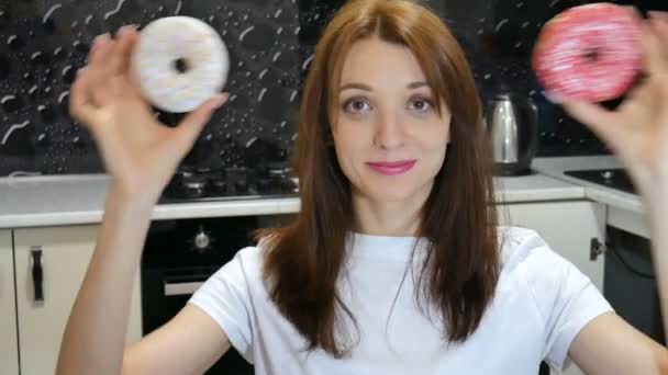 Primer plano retrato de chica divertida con el pelo largo divirtiéndose con rosquillas rojas y blancas contra sus ojos en la cocina en casa — Vídeo de stock