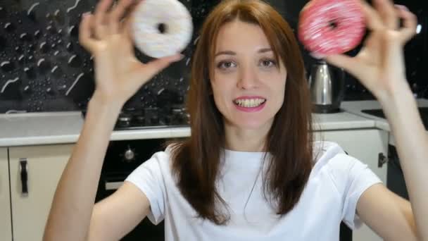 Retrato interior de chica divertida con pelo largo divirtiéndose con rosquillas rojas y blancas contra sus ojos en la cocina en casa — Vídeos de Stock