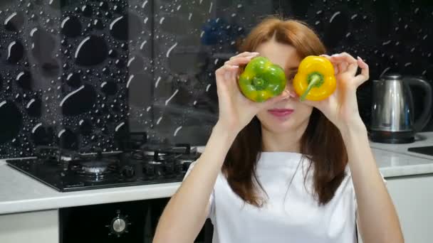 Menina adulta jovem brincando com pimentas amarelas e verdes cobrindo seus olhos com eles antes de preparar comida na cozinha. Estilo de vida saudável e conceito de dieta — Vídeo de Stock