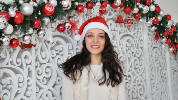 Retrato de una joven muy sonriente en sombrero de santa roja mirando a la cámara en el estudio decorado con Navidad — Vídeo de stock