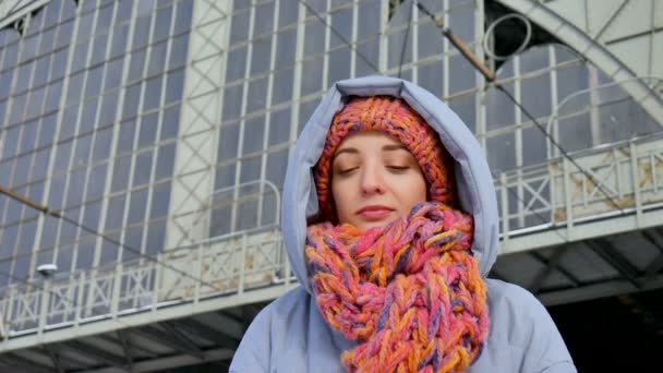 Attraktiv vuxen flicka i blå rock och färgglada mössa med halsduk leende medan du väntar på stationen under solig kall dag. — Stockvideo