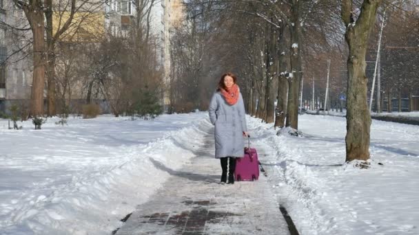 Junge attraktive Frau in warmem Mantel und buntem Strickmuster, die ihren großen violetten Koffer im Vorfrühling auf blauem Himmelshintergrund im Freien zieht — Stockvideo