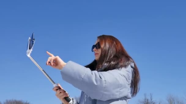 Atractiva mujer morena en gafas de sol haciendo selfie usando smartphone y palo mientras pasa tiempo al aire libre durante el día de primavera en el fondo azul del cielo — Vídeo de stock