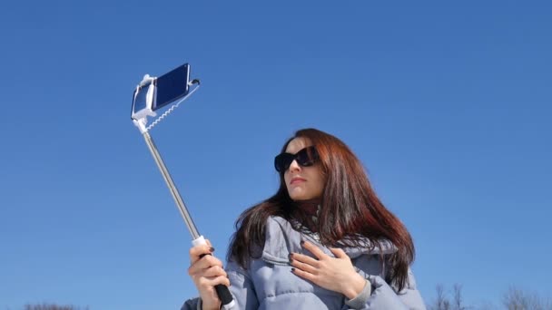 Усміхнена молода жінка в сонцезахисних окулярах фотографує себе, використовуючи смартфон і палицю селфі в парку під час сонячного дня ранньої весни на фоні блакитного неба — стокове відео