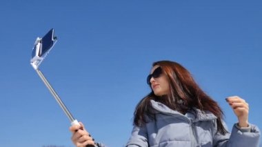Mavi gökyüzü arka planda güneşli bahar gününde onu akıllı telefon ve sopa kullanarak parkta bir selfie çekerken poz karanlık güneş gözlüğü inanılmaz yetişkin kız