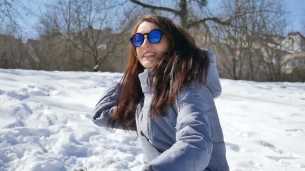ブルーミラーサングラスとストレートダークヘアの素晴らしい大人の女の子は晴れているが、寒い日の午前中に公園で誰かと一緒に遊んで雪玉を投げる。ウィンターゲームコンセプト — ストック動画