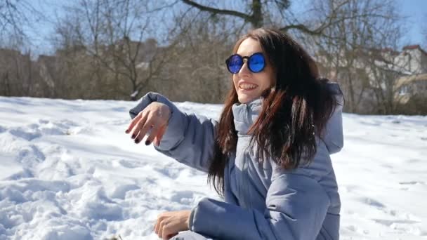 美丽的黑发在蓝色镜像太阳镜和直发投掷雪球扔雪球与人在户外玩在公园里的早晨, 在阳光明媚, 但寒冷的一天在早春 — 图库视频影像