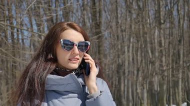 Güneşli sıcak bir gün boyunca ağaçların arka plan üzerinde parkta onun akıllı telefon açık kullanarak biriyle konuşurken genç kadının erken bahar portre