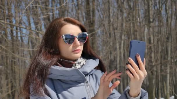 Aantrekkelijk brunette meisje in modieuze sunglesses fotograferen zichzelf met behulp van blauwe smartphone buiten in het Park op bomen achtergrond tijdens zonnige lente dag — Stockvideo