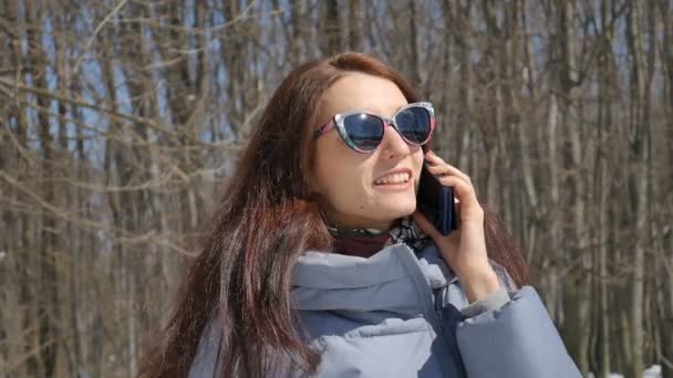 Chica morena atractiva en gafas de sol de moda llamando a alguien que usa el teléfono inteligente azul al aire libre en el parque en el fondo de los árboles durante el soleado día de primavera — Vídeos de Stock