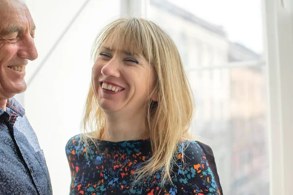 Loira bonita que está perto da janela com seu marido sênior e sorrindo. Conceito de Diferença de Idade . — Fotografia de Stock