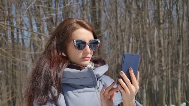 Menina morena atraente em sunglesses na moda chamando alguém usando o smartphone azul ao ar livre no parque no fundo das árvores durante o dia ensolarado da primavera — Vídeo de Stock