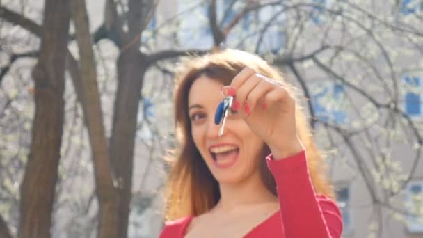 Feliz mujer sonriente sosteniendo su nuevo apartamento o llaves del coche en una mano con manicura roja al aire libre durante la primavera en el fondo urbano. Centrarse en la clave . — Vídeo de stock