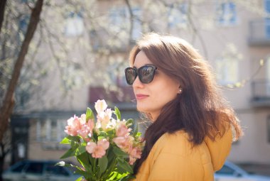 Güneş parlıyor iken güneşli bahar gününde renkli çiçekler büyük bir buket ile açık havada yürüyen güneş gözlüğü şık çekici esmer kadın Outdoors portre