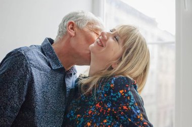 Hot and Sexy Middle-aged Woman Enjoying Kissing of Her Elderly Husband Standing near Opened Window inside Their Home. Couple with Age Difference clipart