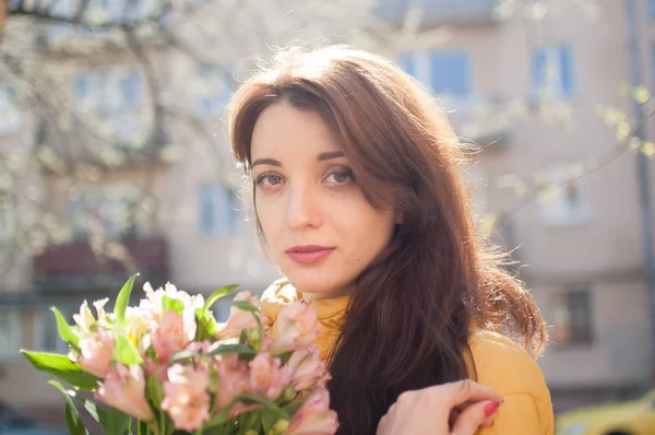 Retrato de mujer joven atractiva y hermosa en chaqueta amarilla sosteniendo un gran ramo de flores de colores al aire libre cerca de los edificios en el fondo — Foto de Stock