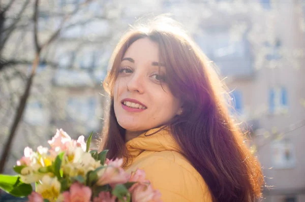Al aire libre retrato femenino de chica morena atractiva en chaqueta amarilla sosteniendo un gran ramo de flores de colores disfrutando de la primavera y mirando a la cámara tne en el fondo del edificio —  Fotos de Stock