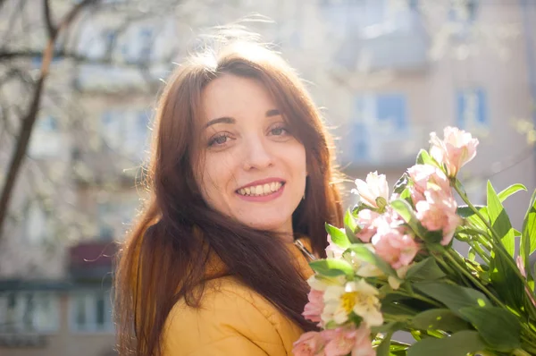 Female portrait of sensual brunette woman in yellow jacket holding a big bouquet of colorful flowers and looking at tne camera on building background during early spring time — Stock Photo, Image