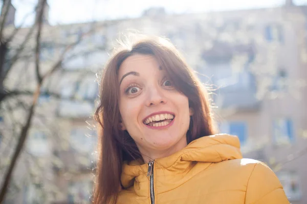 Эмоциональная девушка в ярко-желтой куртке улыбается и смотрит в камеру в раннее весеннее время на открытом воздухе — стоковое фото