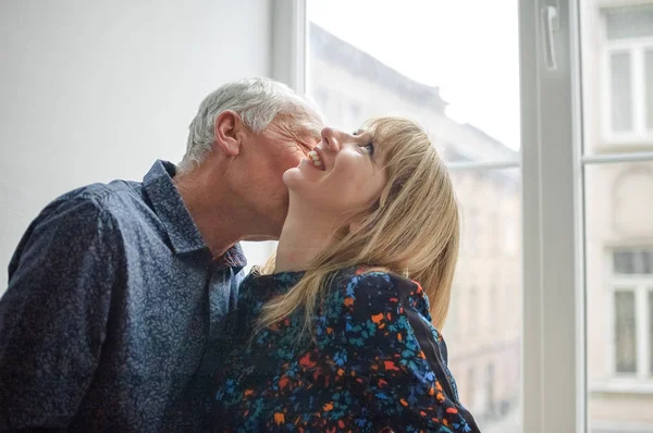 Preciosa pareja con diferencia de edad besándose cerca de la ventana abierta dentro de la casa durante la primavera . —  Fotos de Stock