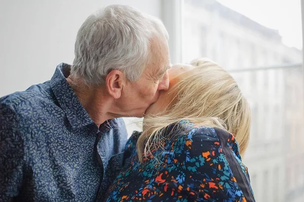 Caliente y sexy mujer de mediana edad disfrutando de besar a su marido mayor de pie cerca de la ventana abierta dentro de su hogar. Pareja con diferencia de edad —  Fotos de Stock