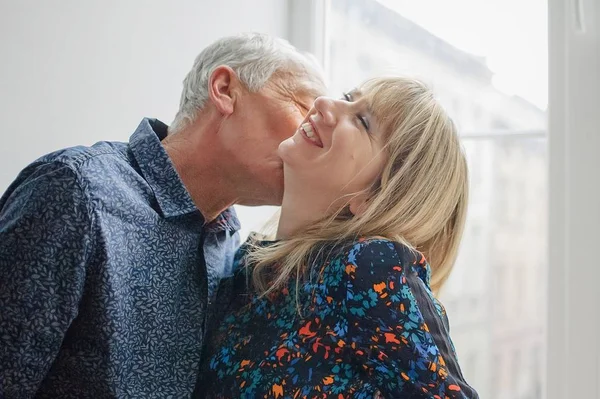 Caliente y sexy mujer de mediana edad disfrutando de besar a su marido mayor de pie cerca de la ventana abierta dentro de su hogar. Pareja con diferencia de edad —  Fotos de Stock