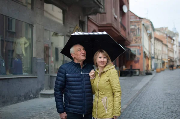 Yaşlı adam ve onun genç sarı saçlı eşi asfalt sokakta şemsiyelerinin altında ayakta birbirlerine kucaklayan açık hava portresi. Yaş farkı olan çift. — Stok fotoğraf