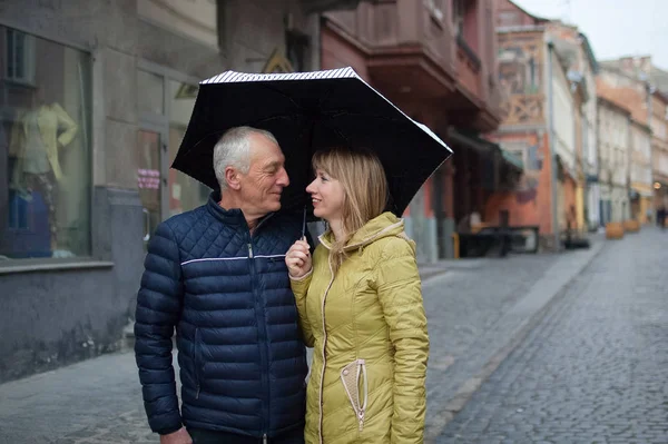 Yaş farkı olan romantik çift, asfalt sokakta şemsiyelerinin altında durarak ve gülümseyerek birbirlerine bakıyorlar.. — Stok fotoğraf