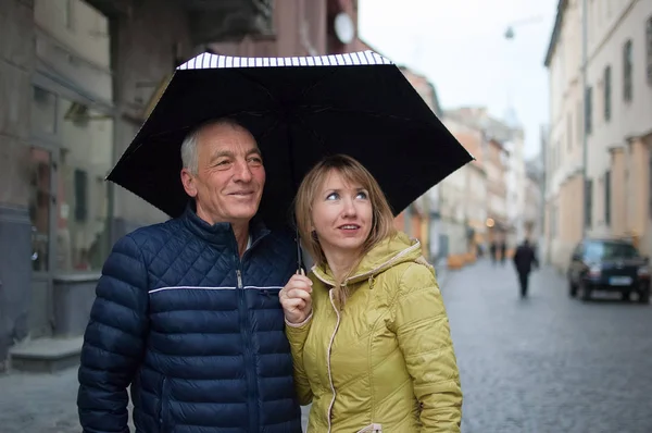 Крупный план портрета пожилого человека и его молодой белокурой жены, обнимающей друг друга и стоящей под зонтиком на асфальтированной улице. Пара с разницей в возрасте . — стоковое фото