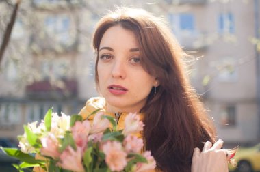 Sarı ceketli şehvetli esmer kadın kadın portresi renkli çiçekler büyük bir buket tutan ve erken bahar aylarında bina arka plan üzerinde tne kamera bakarak