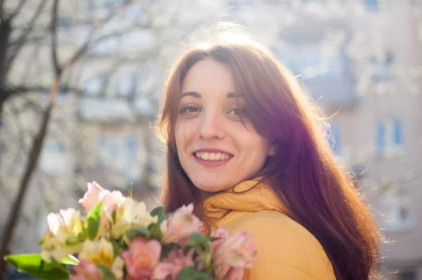 Al aire libre retrato femenino de chica morena atractiva en chaqueta amarilla sosteniendo un gran ramo de flores de colores disfrutando de la primavera y mirando a la cámara tne en el fondo del edificio —  Fotos de Stock