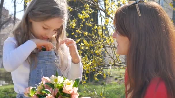 Kleine blonde dochter neemt een bloem uit het boeket en zet hem in een zak van haar denim in het algemeen. Gelukkige moeders dag. Familie vakantie en saamhorigheid. — Stockvideo