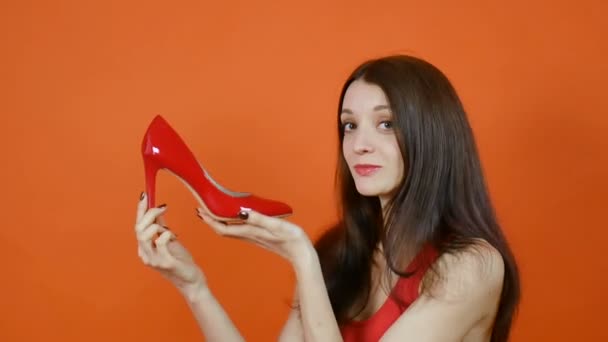 Ein schönes junges braunhaariges Mädchen untersucht ein Paar rote Schuhe. Emotionen. Kunstporträt in einem Atelier auf orangefarbenem Hintergrund — Stockvideo