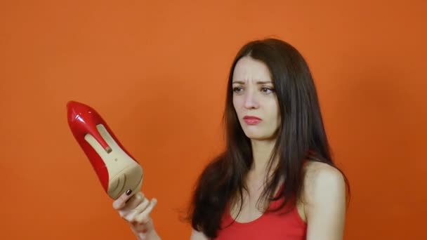 Ein schönes junges braunhaariges Mädchen untersucht ein Paar rote Schuhe. Emotionen. Kunstporträt in einem Atelier auf orangefarbenem Hintergrund — Stockvideo