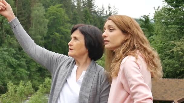 Két nő, fiatal és idős, keresi a távolságot, és beszélnek a háttérben a hegyek és erdők. Hálózati marketing, női barátság, anyák napja, anya és lánya — Stock videók