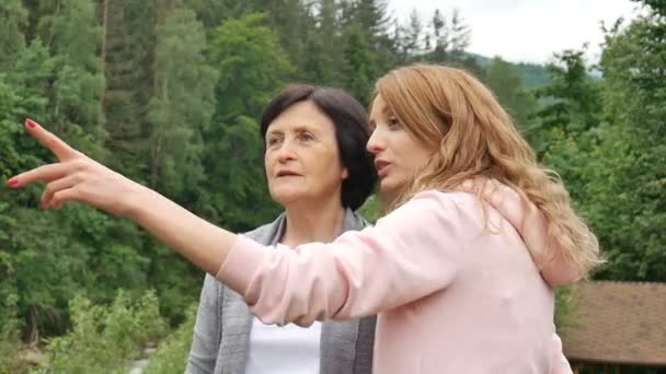 Egy idősebb nyugdíjas anya és felnőtt lánya beszél a háttérben az erdős hegyek. Üdülés a hegyekben, anyák napja, két generáció — Stock videók