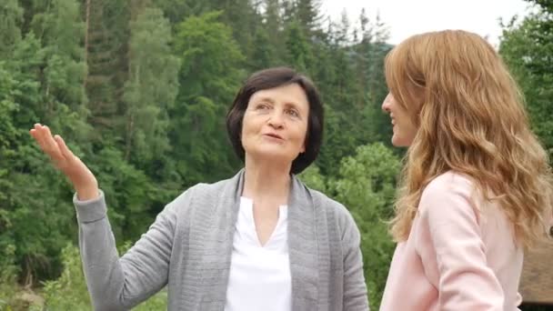 Две женщины, молодые и пожилые, смотрят вдаль и разговаривают на фоне гор и лесов. Сетевой маркетинг, женская дружба, день матери, мать и дочь — стоковое видео