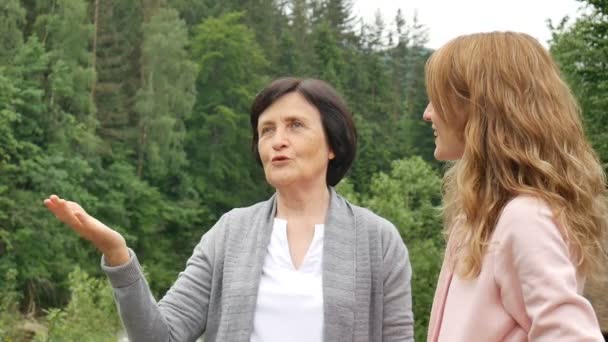 Två kvinnor, unga och äldre, tittar in i fjärran och pratar mot bakgrund av berg och skog. Nätverksmarknadsföring, kvinnlig vänskap, mors dag, mor och dotter — Stockvideo