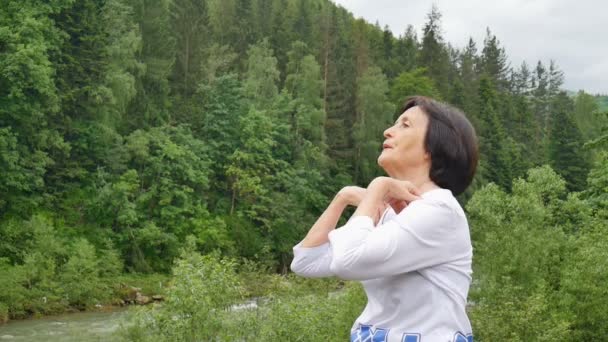 Старшая женщина с короткими темными волосами делает растяжку упражнения для отдыха утром на улице над ландшафтом леса и гор — стоковое видео