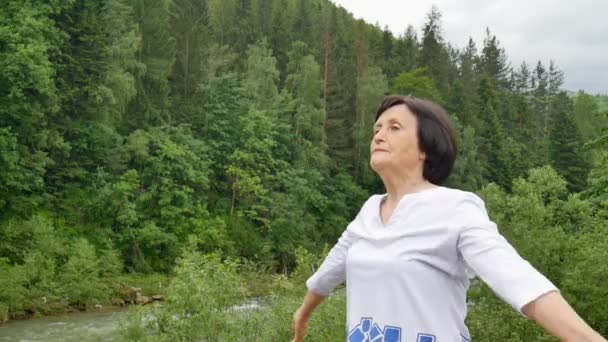 Senior vrouw doet een stretching oefening voor de bovenarmen buiten over landschap van bos en bergen — Stockvideo
